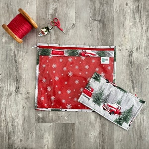 Pode incluir: Duas bolsas transparentes com z&iacute;per com padr&otilde;es de flocos de neve vermelhos e brancos e uma cena de inverno com um caminh&atilde;o vermelho e pinheiros. A bolsa maior tem uma etiqueta verde com o texto "Sewing Shop".