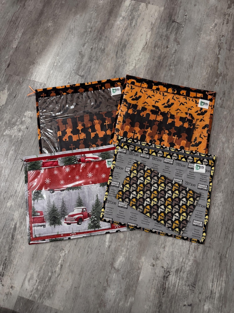 Pode incluir: Quatro bolsas com z&iacute;per diferentes com v&aacute;rios designs. As bolsas s&atilde;o feitas de tecido e t&ecirc;m uma janela de pl&aacute;stico transparente. Os designs incluem uma &aacute;rvore de Natal e um caminh&atilde;o, um cemit&eacute;rio com morcegos, um padr&atilde;o de trabalhadores de escrit&oacute;rio e um padr&atilde;o de trabalhadores de escrit&oacute;rio com o texto "Dunder Mifflin" repetido.