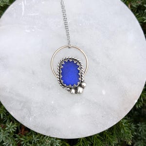 May include: A silver chain necklace with a blue glass pendant set in a silver frame. The pendant is surrounded by a gold circle.