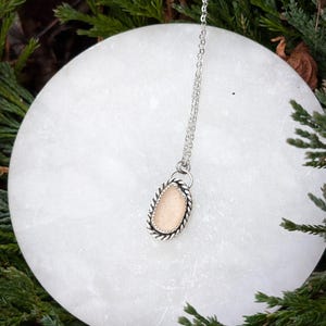 May include: A silver chain necklace with a delicate pendant featuring a light brown oval-shaped stone set in a silver frame with a rope-like design.