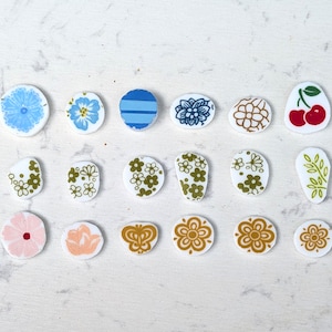 May include: A collection of 16 round ceramic tiles with various floral and geometric patterns. The tiles are white with blue, green, and yellow accents. Some tiles feature cherries, butterflies, and leaves.