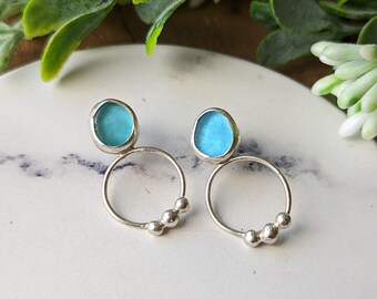 May include: A pair of silver earrings with a small, round, light blue glass stone on each earring. The earrings have a small circle with three silver beads on each side of the stone.
