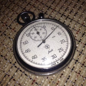 May include: A vintage stopwatch with a silver casing and a white face. The face features black numerals and markings for seconds and fractions of a second. The stopwatch has a small dial at the top and the text "MADE IN USSR" is visible.