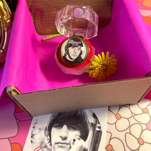 Könnte beinhalten: Eine Ringbox mit rotem Samt-Innenfutter und einem durchsichtigen Plastikdeckel. Der Ring zeigt ein Schwarzweißfoto von Ringo Starr mit dem Wort "Ringo" auf dem Ring. Ein Aufkleber mit dem Text "I Love Ringo" und einem roten Herzen ist ebenfalls enthalten.