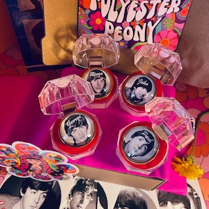 May include: Four clear plastic ring boxes with red velvet interiors. Each box features a different Beatles member: Ringo, John, George, and Paul. The boxes are arranged on a pink and floral patterned surface. The text "Polyester Peony" is visible on a pink and floral patterned coaster.