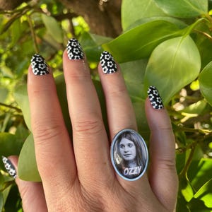 May include: A silver-toned ring with an oval face featuring a black and white image of a person with the word "OZZY" in white. The nails are painted with a black and white floral pattern. The background is green foliage.