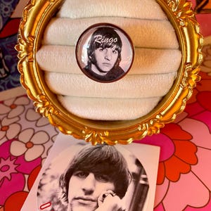Könnte beinhalten: Ein silberner Ring mit einem Glas-Cameo, das ein Schwarzweißfoto von Ringo Starr zeigt. Das Cameo trägt den Schriftzug "Ringo" in weißer, kursiv geschriebener Schrift. Der Ring wird auf einem weißen Ringhalter in einem goldenen Rahmen präsentiert. Unter dem Ring befindet sich ein Aufkleber mit einem Schwarzweißfoto von Ringo Starr. Der Aufkleber trägt den Schriftzug "I Love Ringo" in roten Buchstaben und ein rotes Herz.