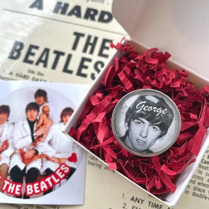 May include: A white box filled with red tissue paper. Inside the box is a round, silver pinback button featuring a black and white photo of George Harrison from the Beatles. The button has the word "George" printed in black letters at the top. The box is resting on a white piece of paper with the words "Songs from the film. A HARD DAY'S NIGHT THE BEATLES" printed in black letters.