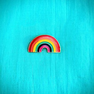 May include: A rainbow-colored enamel pin with a gold outline. The pin is shaped like a rainbow arch with seven colors: red, orange, yellow, green, blue, indigo, and violet.