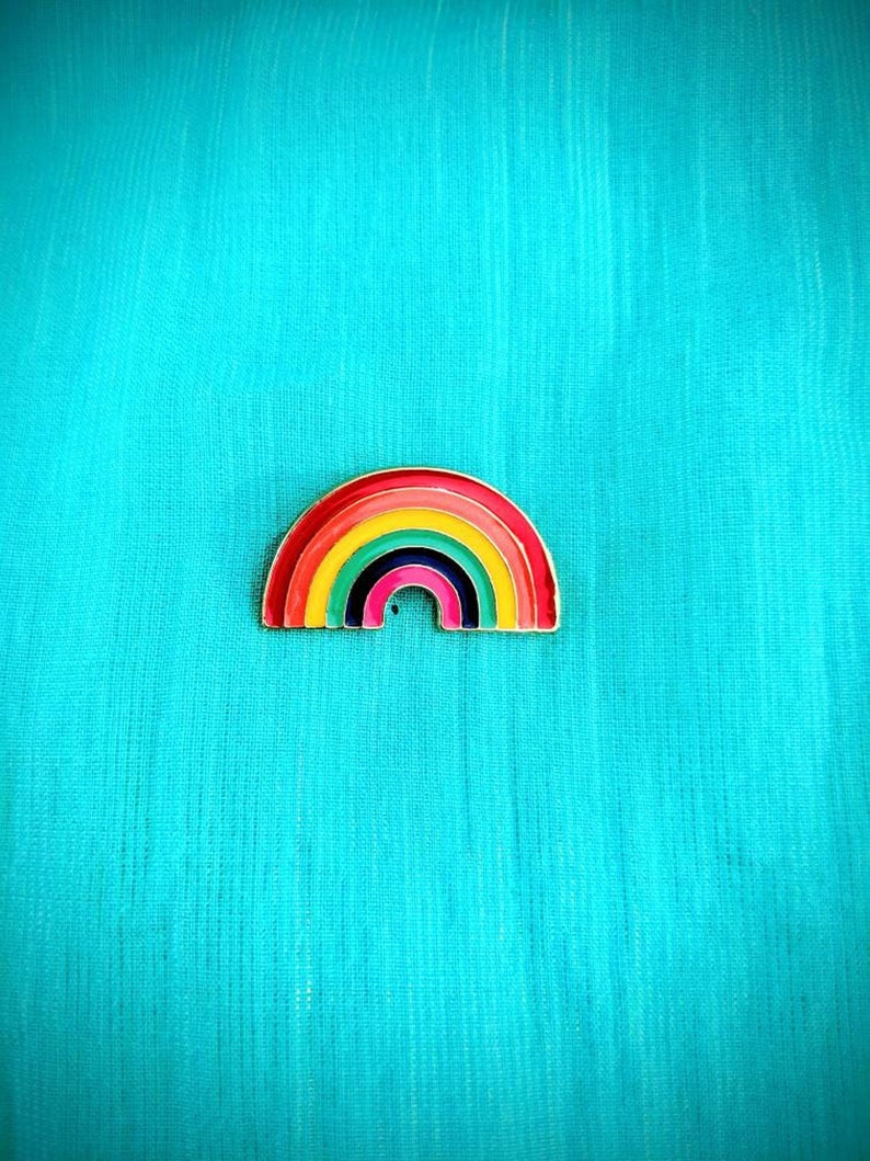 May include: A rainbow-colored enamel pin with a gold outline. The pin is shaped like a rainbow arch with seven colors: red, orange, yellow, green, blue, indigo, and violet.