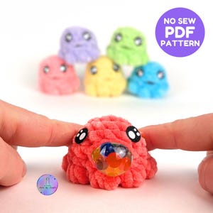 Puede incluir: Un juguete de peluche de color rojo brillante tejido a crochet con una bola de plástico transparente llena de cuentas de colores. El juguete tiene dos ojos negros y una boca redonda y esponjosa. El juguete está siendo sostenido por dos manos. El texto "NO SEW PDF PATTERN" está en un círculo morado en la esquina superior derecha de la imagen.