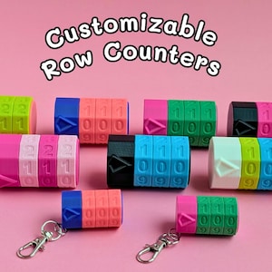 May include: A collection of colourful, customizable row counters in shades of pink, green, blue, and black. Each counter shows numbers and has a rotating mechanism. Some have a keychain. The text "Customizable Row Counters" is displayed at the top.