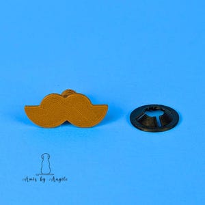 May include: A golden-brown moustache-shaped item and a black, circular object with a slit, against a bright blue backdrop. The moustache has a textured surface. The items appear to be decorative or functional.