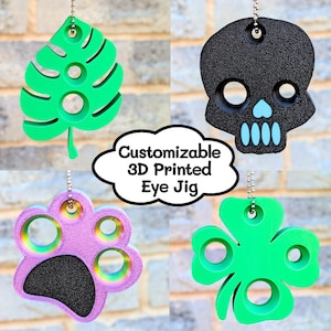 May include: A selection of 3D-printed eye jigs, including a green leaf, a black skull with blue details, a purple paw print, and a green four-leaf clover. Each is suspended by a chain. The text "Customizable 3D Printed Eye Jig" is visible.