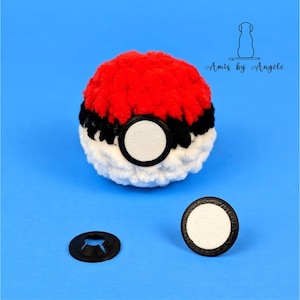 May include: A red, black and white crocheted Pokeball with a white felt circle in the centre. The Pokeball is sitting on a blue surface with two black plastic rings and a black plastic ring with three prongs.