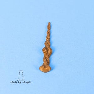 May include: A brown, twisted wand-like object with a rounded base, set against a light blue background. The wand has a detailed, layered design, resembling wood grain. The image also includes a logo with a dog silhouette and the text "Amis by Angele."