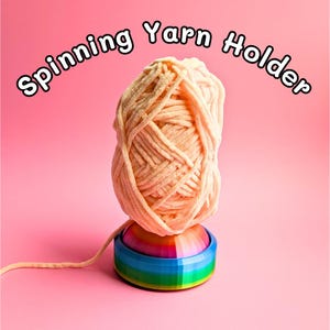 May include: A ball of peach-coloured yarn is displayed on a rainbow-coloured spinning yarn holder. The holder has a blue, green, and yellow base. The words "Spinning Yarn Holder" are arched above the yarn ball.