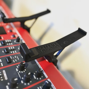 Puede incluir: Un soporte de tel&eacute;fono de pl&aacute;stico negro con la palabra "YAMAHA" impresa en &eacute;l. El soporte est&aacute; unido a un teclado rojo.