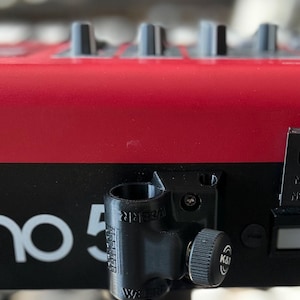 May include: A black plastic cylindrical mount attached to a red and black electronic keyboard. The mount features a textured surface with the text "WEBRR" embossed on the side and a circular adjustment knob labelled with the "K&M" logo.