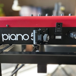 May include: A red and black digital keyboard with the text piano 5 printed in white on the front panel. The device features a black mounting bracket with a circular knob attached to the centre, along with a power input port and a cable.