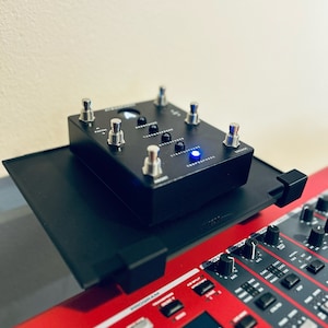 May include: A black foot pedal with five silver buttons and a blue light. The pedal is sitting on top of a red keyboard with black buttons and knobs.