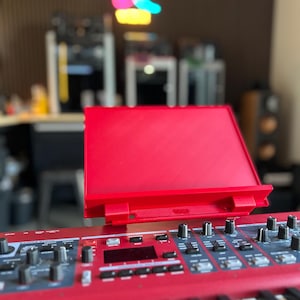 May include: A bright red music stand attachment mounted on a red electronic keyboard. The stand features a flat rectangular back panel with a horizontal ledge at the base, designed to hold sheet music or a tablet above the control panel of the instrument.