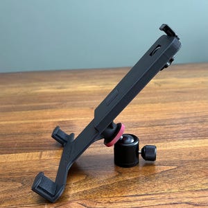 Könnte beinhalten: Ein schwarzer Tablethalter mit rotem Akzent an einem schwarzen Kugelkopf. Der Halter ist abgewinkelt, mit einer Klemme oben und zwei Stützarmen unten. Das Gerät befindet sich auf einer Holzoberfläche.