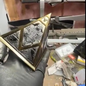 May include: A metallic, geometric object with intricate engraved patterns. The object has a gold-coloured trim and a silver-coloured centre. The design appears to be a decorative piece or a component of a larger structure.