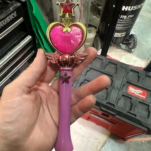 Könnte beinhalten: Ein rosa-goldener Fantasy-Zauberstab mit herzförmigem Edelstein, Sternakzenten und Flügeldarstellungen. Der Zauberstab hat einen langen, schlanken Griff und ist wahrscheinlich ein Cosplay- oder Sammlerstück.