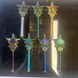 Op de afbeelding: Collectie van zeven stervormige toverstokken met gouden accenten. Elke toverstaf heeft een verschillend gekleurde handgreep en een symbool in het midden van de ster. De toverstokken hebben een kroonontwerp aan de basis van de ster en een zilveren, turquoise, oranje, paarse, blauwe en groene handgreep.