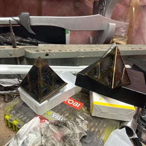May include: Two bronze pyramid-shaped objects with detailed patterns. One rests on a white plastic tray, the other on a black box. A silver sword-like item is in the background. The items are on a cluttered workbench.