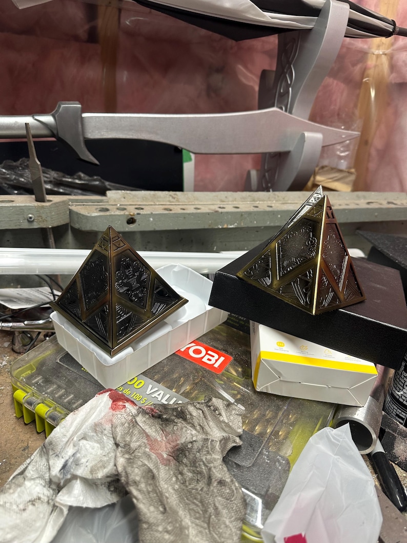 May include: Two bronze pyramid-shaped objects with detailed patterns. One rests on a white plastic tray, the other on a black box. A silver sword-like item is in the background. The items are on a cluttered workbench.
