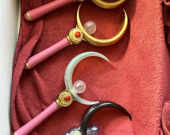 Könnte beinhalten: Vier Halbmond-Zauberstäbe mit rosa und schwarzen Griffen und goldenen Akzenten. Jeder Zauberstab hat eine klare Kugel oben.