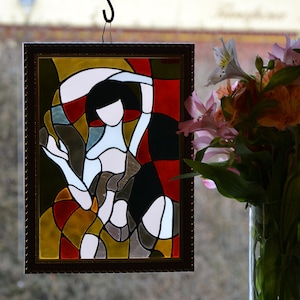 Puede incluir: Una obra de arte de vitral enmarcada que representa una figura estilizada en un patrón geométrico colorido. La figura está en una pose con los brazos levantados, y la obra de arte está sobre un fondo borroso de una ventana y flores.