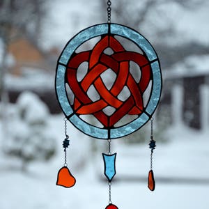 Puede incluir: Un atrapasol de vidrio coloreado con un diseño de nudo celta rojo en un círculo azul. Tiene tres colgantes: un corazón rojo, un corazón naranja y una flecha azul.