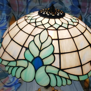May include: A stained glass lamp with a white and green floral design. The lamp has a blue center and a gold base.