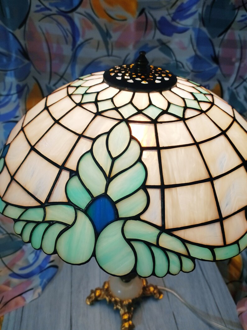 May include: A stained glass lamp with a white and green floral design. The lamp has a blue center and a gold base.