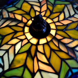 May include: A stained glass lampshade with a peacock feather design. The lampshade is made of colorful glass pieces in shades of blue, green, yellow, and orange. The center of the lampshade features a circular design with a yellow and orange center.