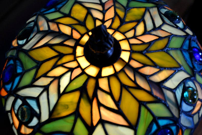 May include: A stained glass lampshade with a peacock feather design. The lampshade is made of colorful glass pieces in shades of blue, green, yellow, and orange. The center of the lampshade features a circular design with a yellow and orange center.