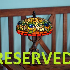 May include: A stained glass table lamp with a peacock feather design. The lamp has a brown base and a colorful glass shade with green, blue, and yellow accents. The lamp is sitting on a wooden table with the word "RESERVED" printed on it.