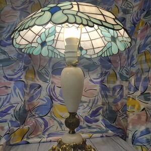 May include: A white alabaster table lamp with a stained glass shade featuring a floral design in shades of blue and green. The lamp has a gold base with ornate details.