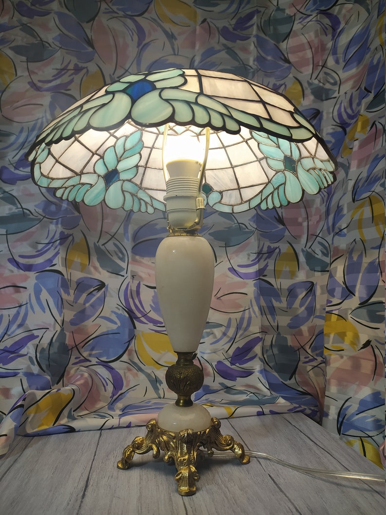 May include: A white alabaster table lamp with a stained glass shade featuring a floral design in shades of blue and green. The lamp has a gold base with ornate details.