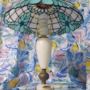 May include: A Tiffany-style table lamp with a white and blue stained glass shade featuring a floral design. The lamp has a white ceramic base with gold accents and a gold-colored metal base.