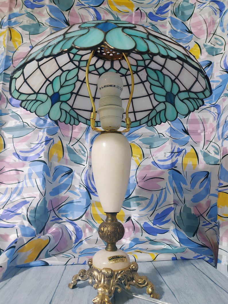 May include: A Tiffany-style table lamp with a white and blue stained glass shade featuring a floral design. The lamp has a white ceramic base with gold accents and a gold-colored metal base.