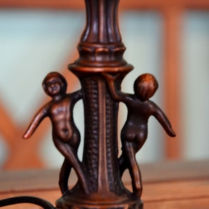 May include: A bronze lamp base with two cherub figures holding onto the base. The lamp base is decorated with intricate details.