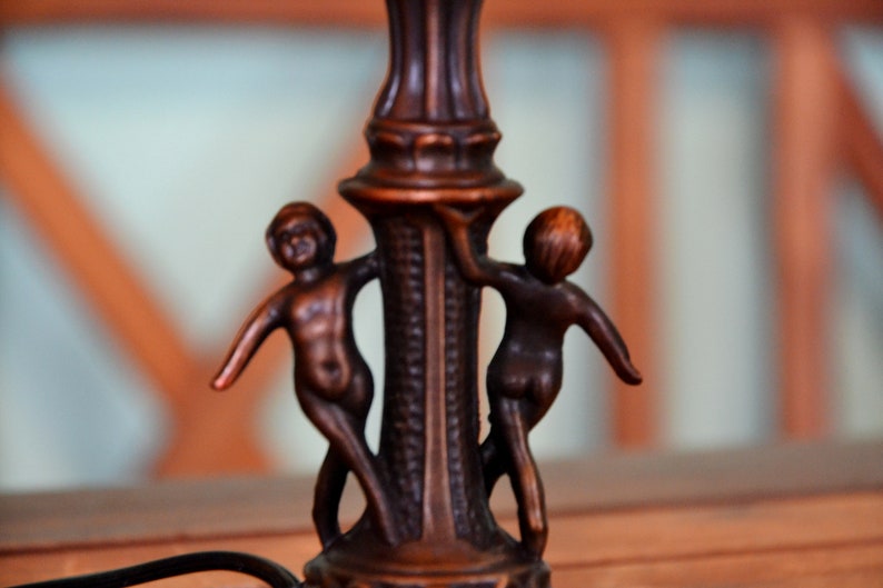 May include: A bronze lamp base with two cherub figures holding onto the base. The lamp base is decorated with intricate details.