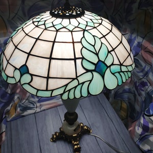 May include: A Tiffany-style table lamp with a white and blue stained glass shade featuring a floral design. The lamp has a gold-tone base with three legs.