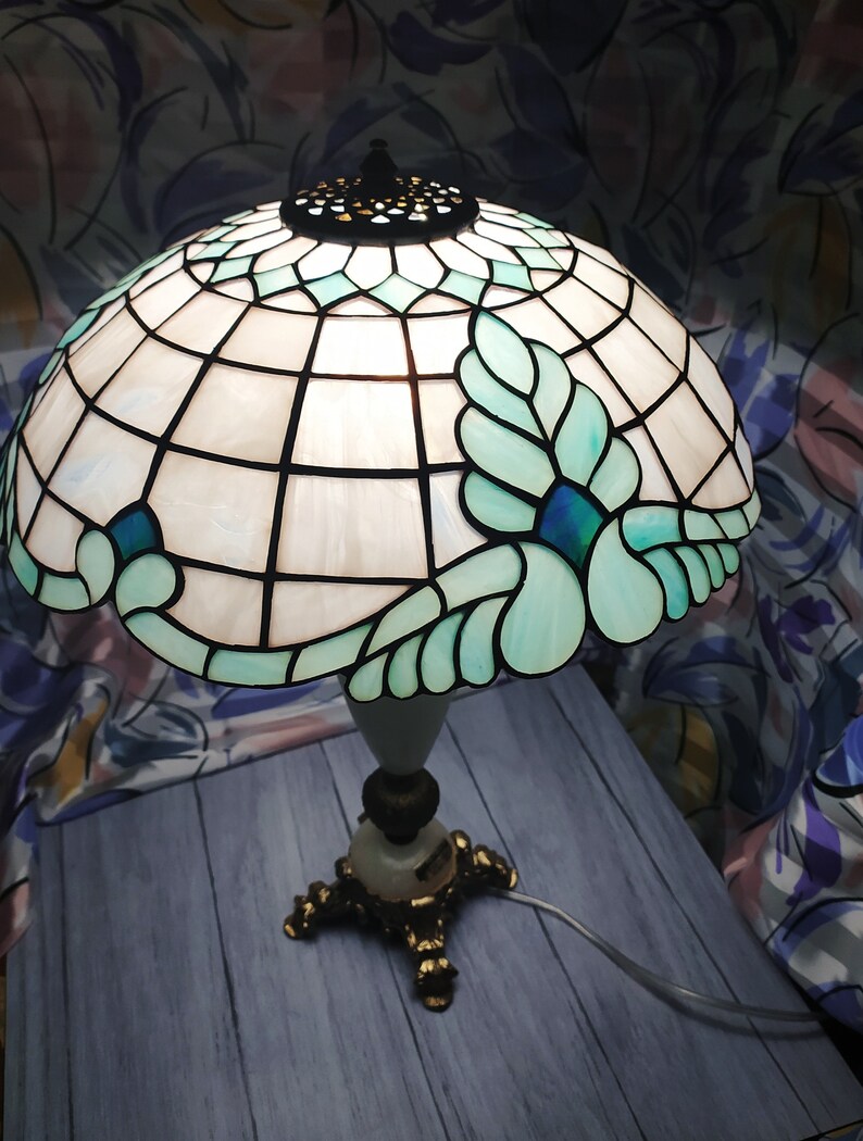 May include: A Tiffany-style table lamp with a white and blue stained glass shade featuring a floral design. The lamp has a gold-tone base with three legs.
