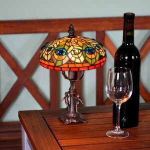 May include: A stained glass table lamp with a peacock design. The lamp has a brown base and a colorful shade with green, blue, and yellow accents. The lamp is lit and casting a warm glow. A glass of wine and a bottle of wine are on the table next to the lamp.