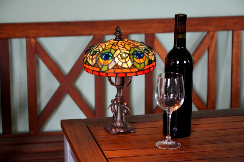May include: A stained glass table lamp with a peacock design. The lamp has a brown base and a colorful shade with green, blue, and yellow accents. The lamp is lit and casting a warm glow. A glass of wine and a bottle of wine are on the table next to the lamp.